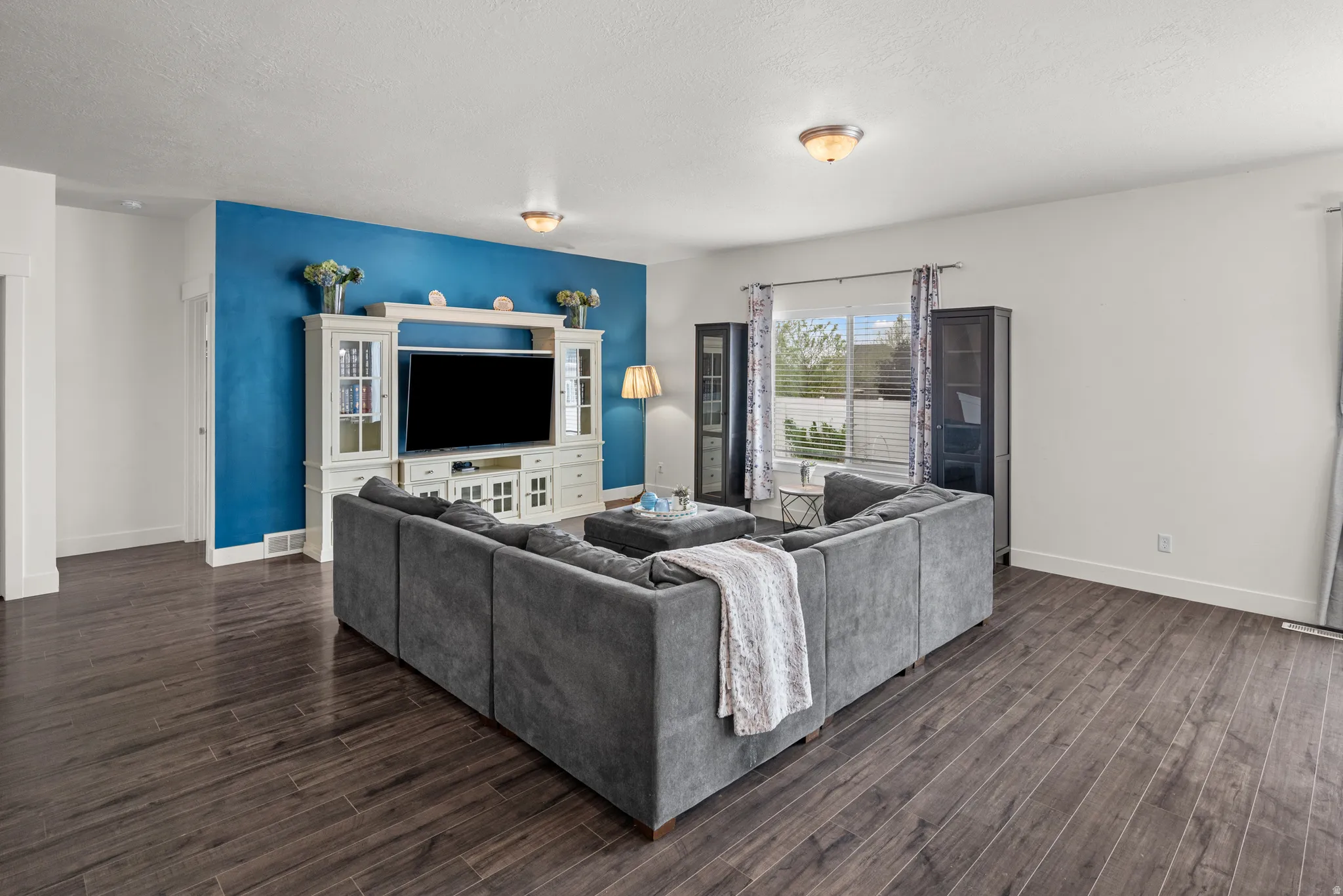 Spacious living area featuring wood-finish flooring, a prominent accent wall, and a large window with blinds