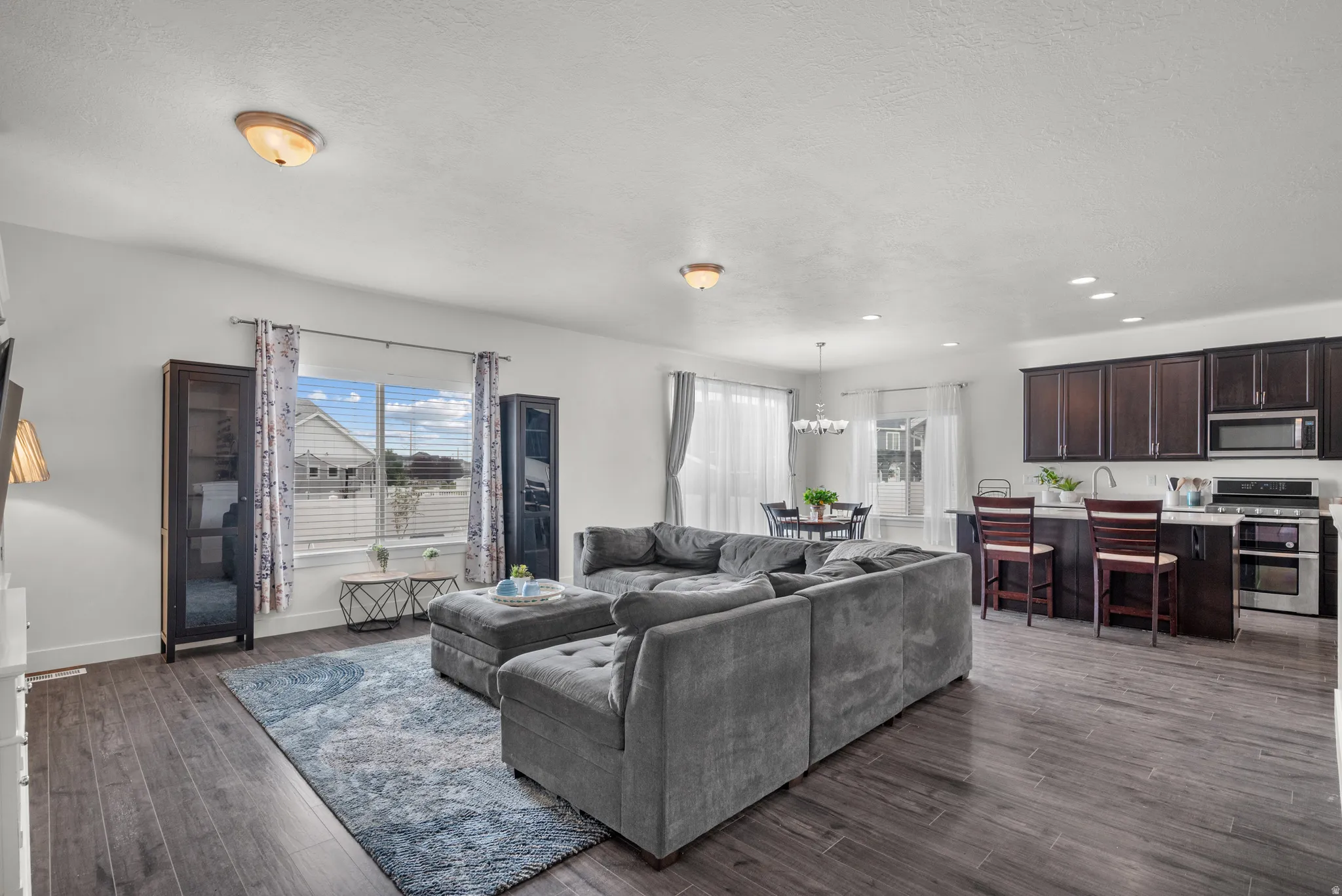Open-concept living area featuring wood-finish flooring, recessed lighting, a modern kitchen with dark cabinetry, stainless steel appliances, and a breakfast bar