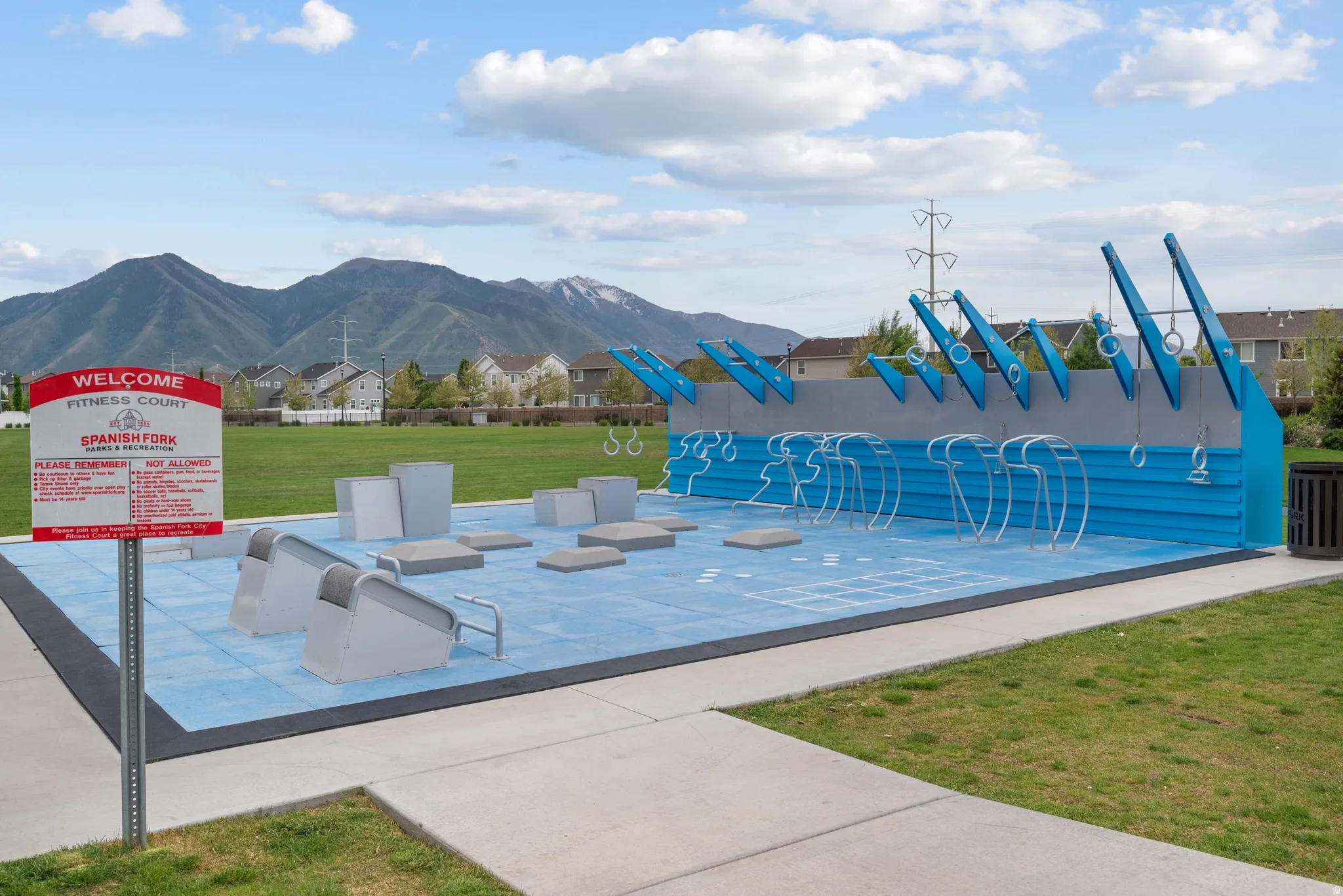 Outdoor fitness court featuring blue and gray padded surfacing, various exercise structures, and surrounding green space at the Legacy Farm Park located directly across the street.