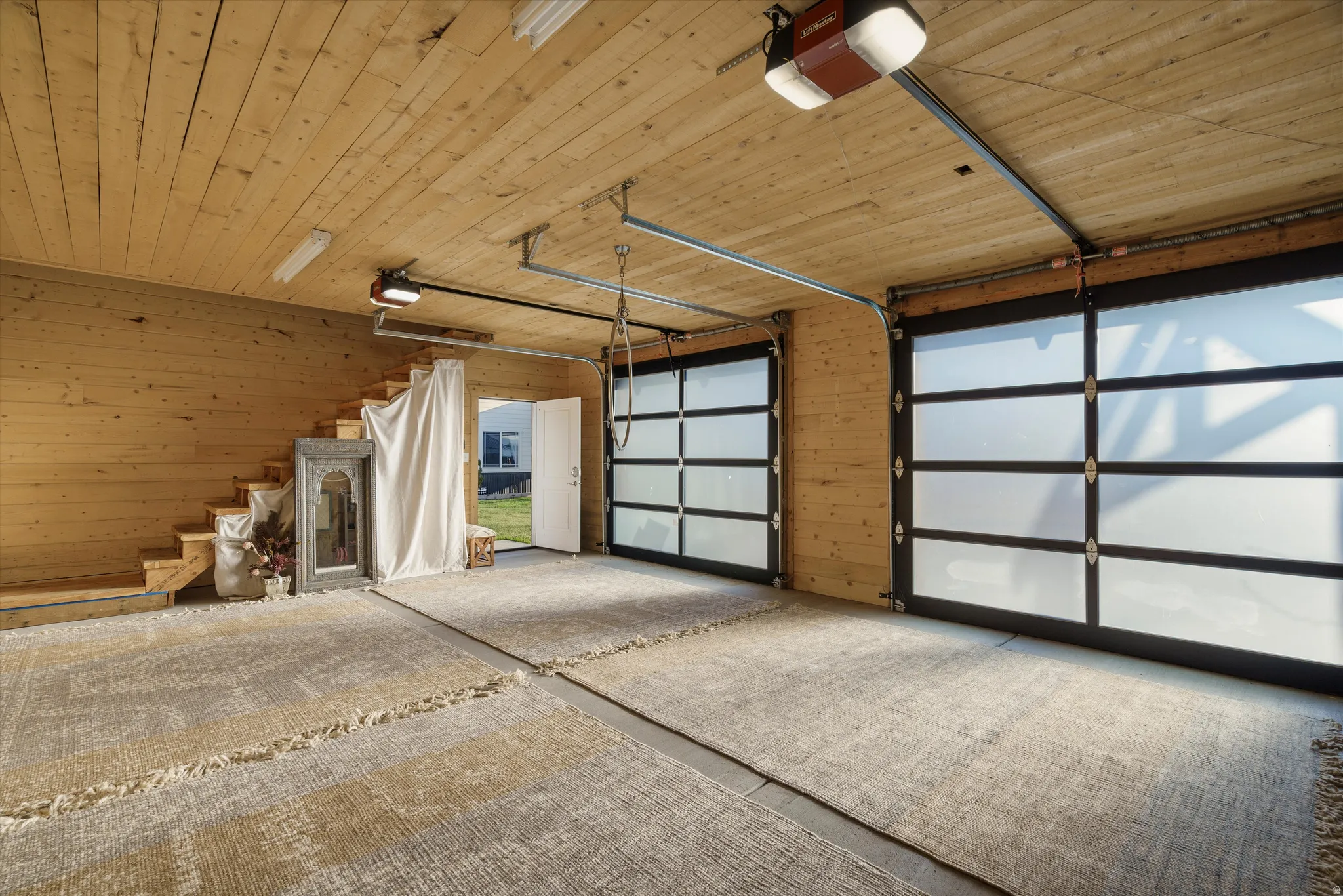 Spacious garage featuring light wood-paneled walls and ceiling, two modern garage doors with frosted glass panels, and a visible staircase leading to an upper level
