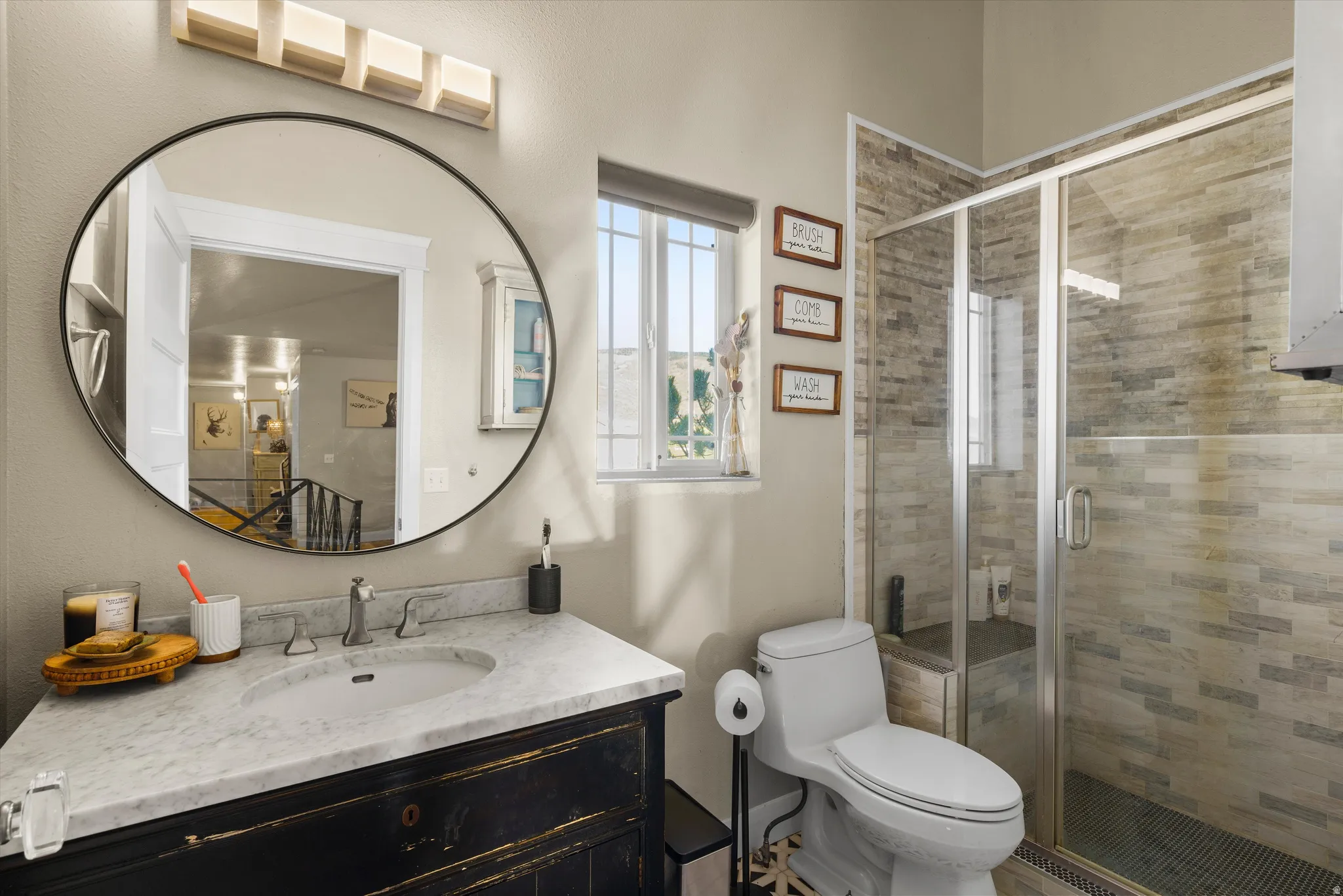 Bathroom featuring a dark wood vanity with a white stone countertop, a round black-framed mirror, and a glass-enclosed shower with stone tile walls
