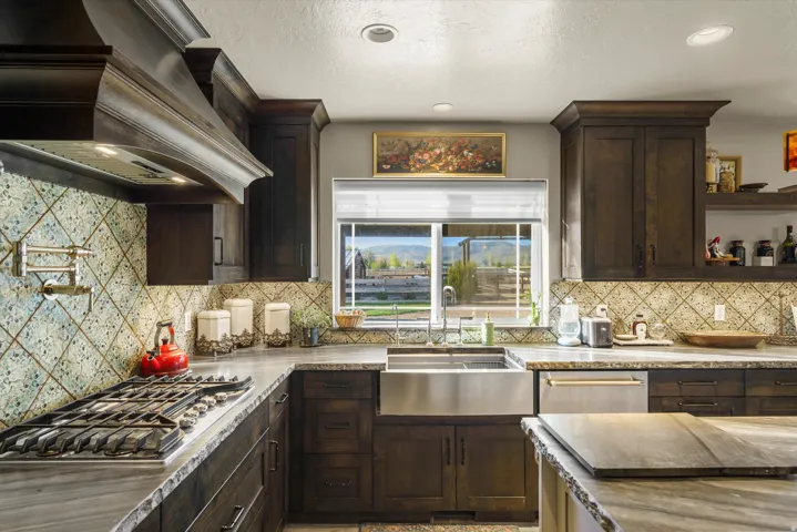Gourmet kitchen featuring dark wood cabinetry, an industrial gas range with pot filler, stainless steel farmhouse sink, integrated dishwasher, and patterned tile backsplash