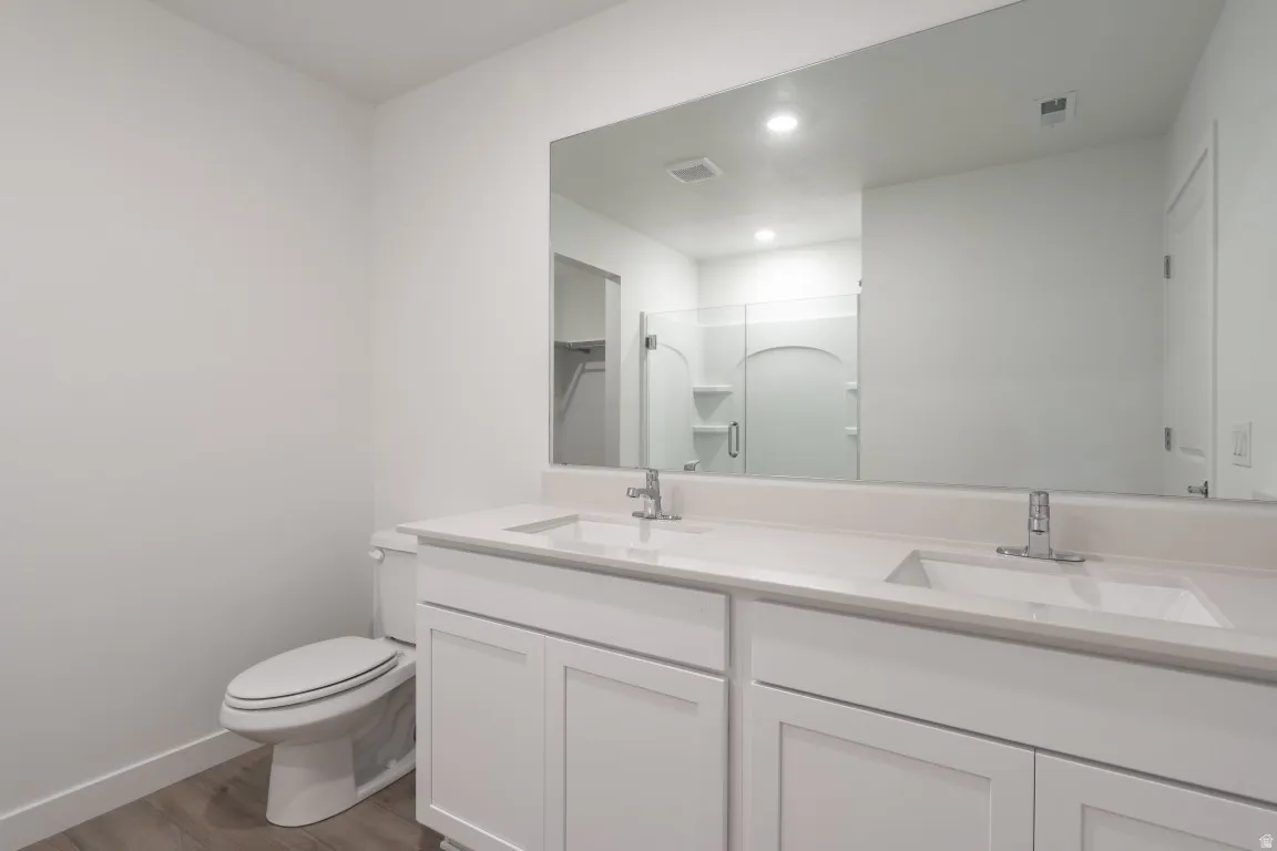 Dual sink vanity with white cabinetry and integrated sinks, wide wall-mounted mirror, wood-finish flooring, and a white toilet