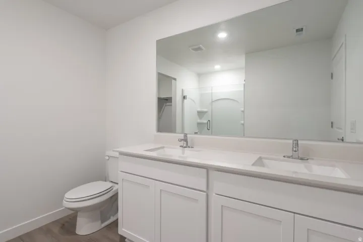 Dual sink vanity with white cabinetry and integrated sinks, wide wall-mounted mirror, wood-finish flooring, and a white toilet