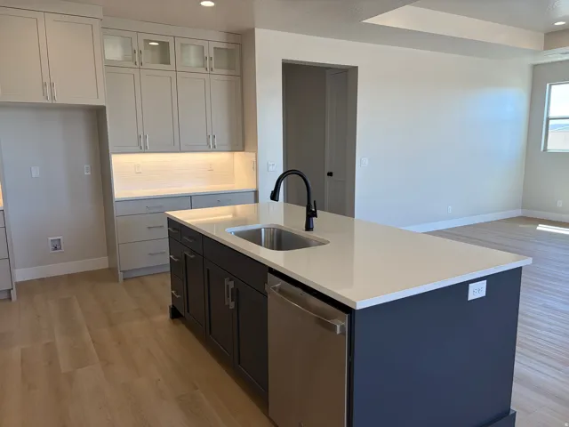 Kitchen island with integrated stainless steel sink and matte black faucet, light wood-finish flooring, white countertops, built-in dishwasher, and recessed lighting