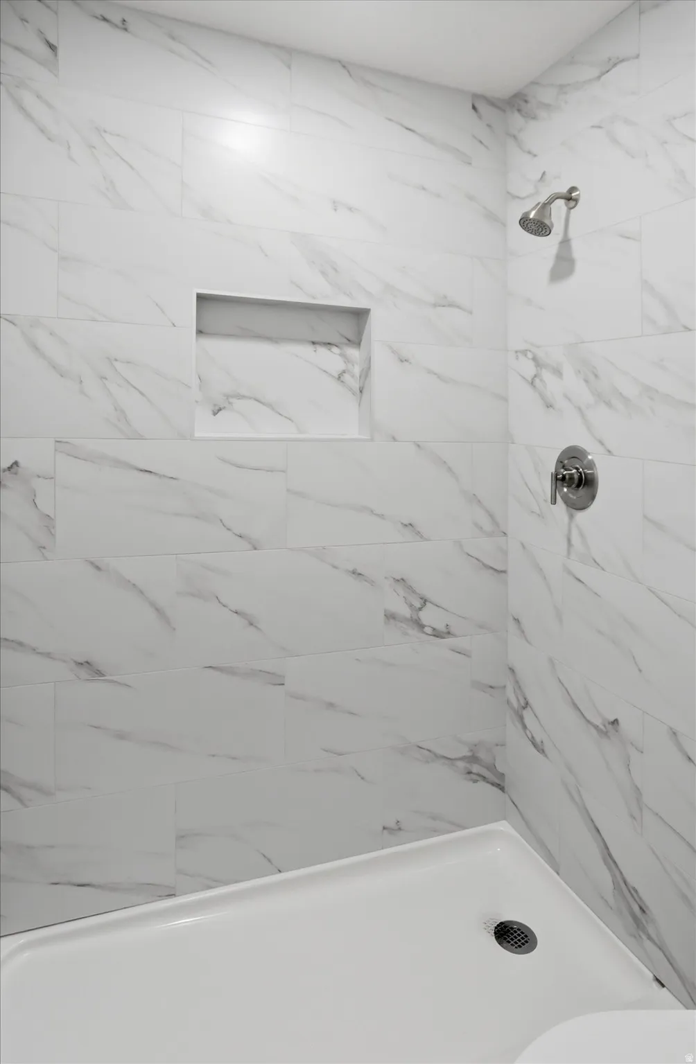 Shower featuring large format white tiled walls with gray veining, a built-in shower niche, and a brushed nickel shower head and control
