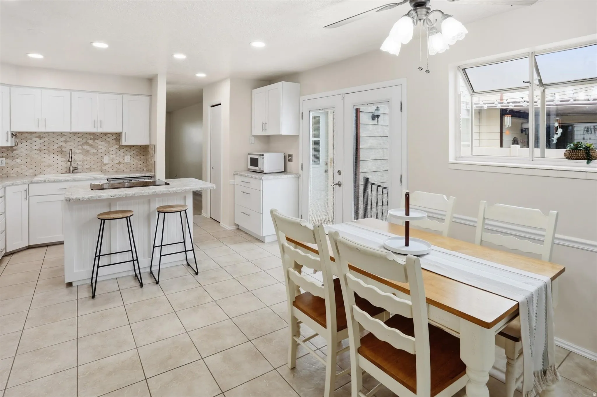 Open-concept kitchen and dining area featuring white shaker cabinetry, light-toned tile flooring, granite-style countertops, and a mosaic tile backsplash