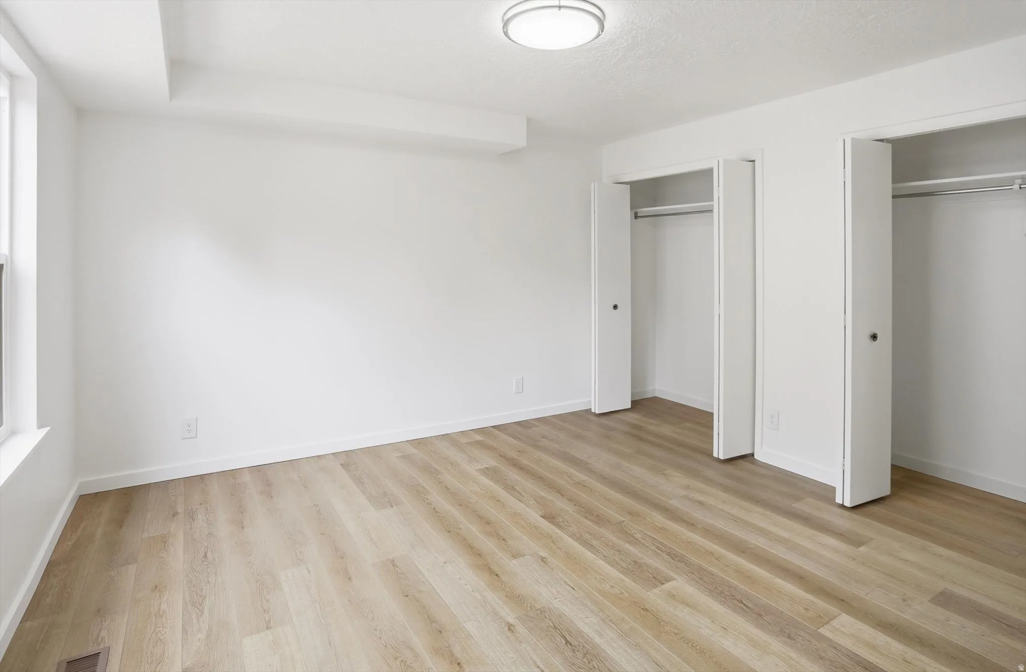 Spacious room featuring light wood-finish flooring, bright white walls, and a modern flush-mount ceiling light