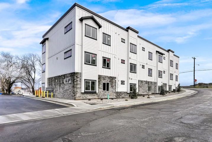 Multi-level building exterior featuring white vertical and horizontal siding with dark trim, stacked stone facade, multiple windows with dark frames, and a paved parking area