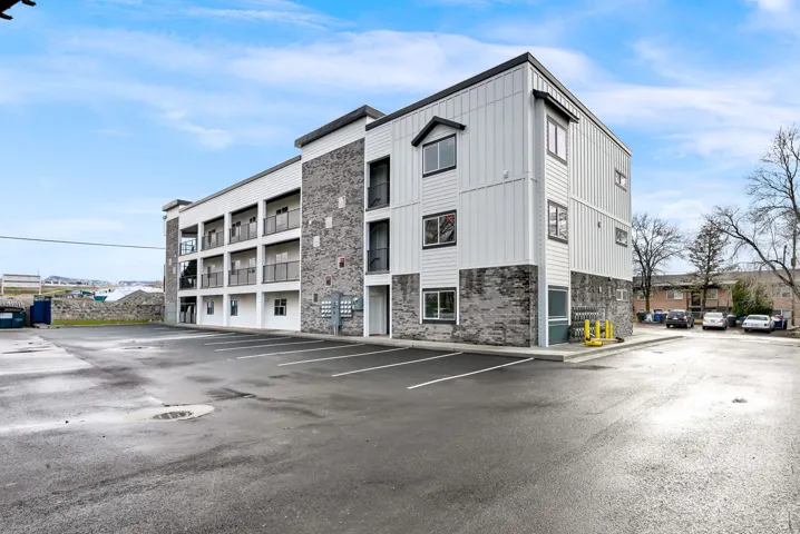 Contemporary multi-story building featuring vertical and horizontal siding panels, stone veneer accents, individual unit balconies with metal railings, and a large paved parking area