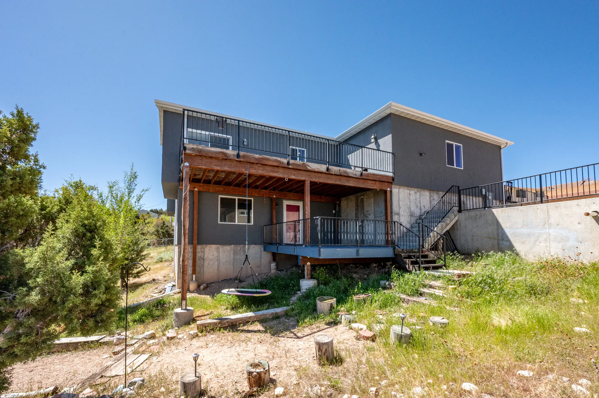 Two-story exterior with a gray facade, featuring an upper-level deck with black railings and a lower-level covered patio with wood supports