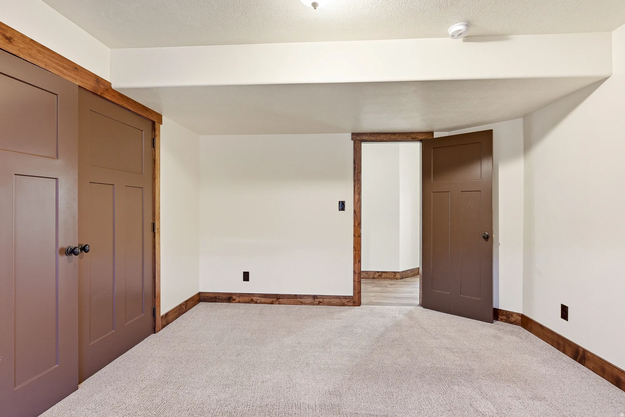 Carpeted room featuring brown trim, two brown doors, and an additional entryway with wood-finish flooring visible beyond