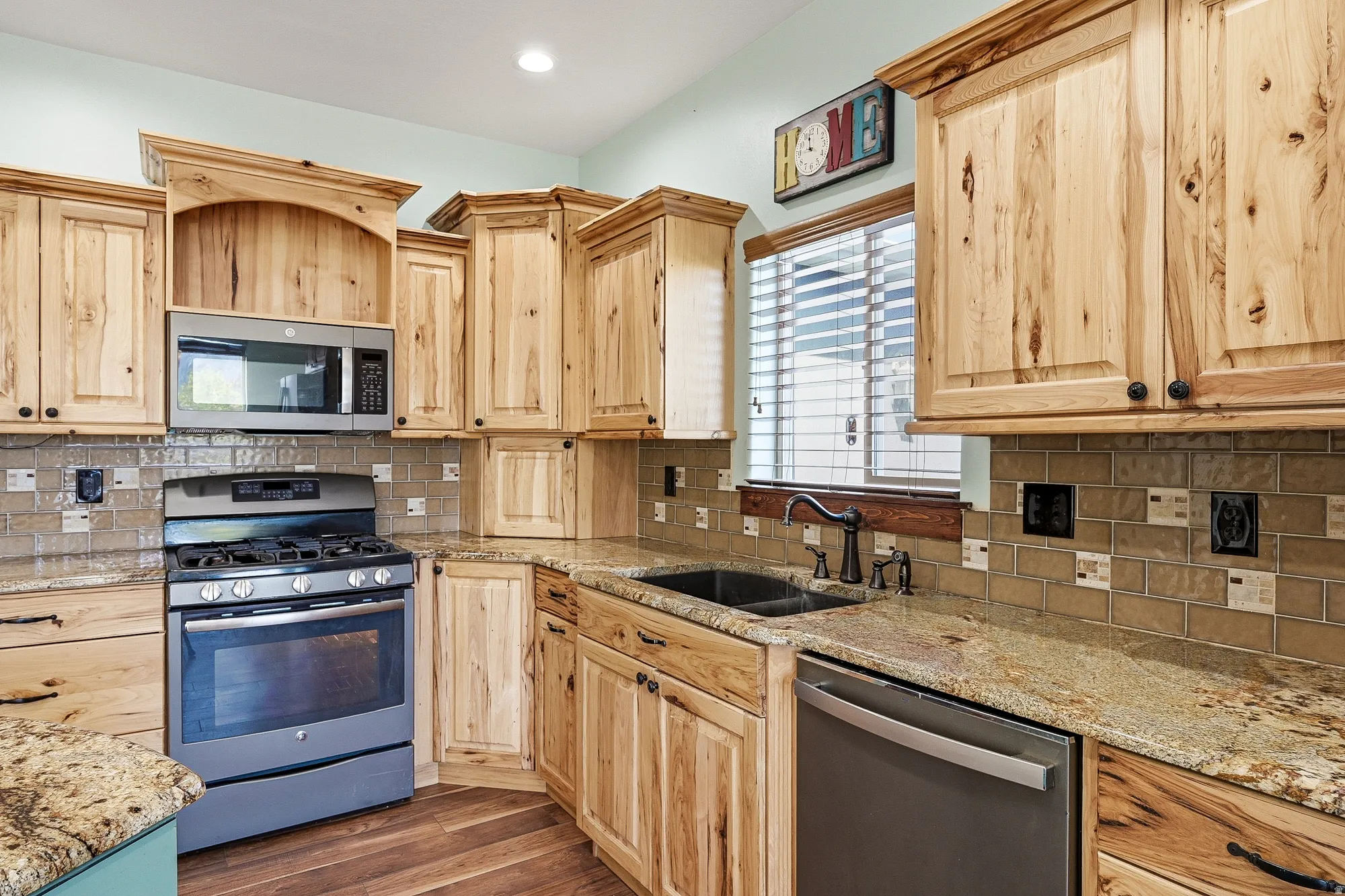 Kitchen featuring natural wood cabinetry, granite countertops, tile backsplash, wood-finish flooring, and stainless steel appliances