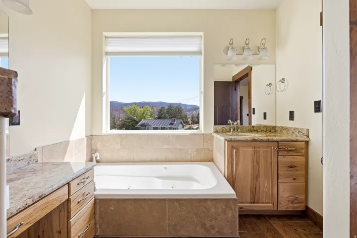 Bathroom featuring a built-in jetted tub with tile surround, a natural wood-finish vanity with a stone countertop and integrated sink, a separate natural wood-finish vanity with a stone countertop, a large window, and wood-finish flooring