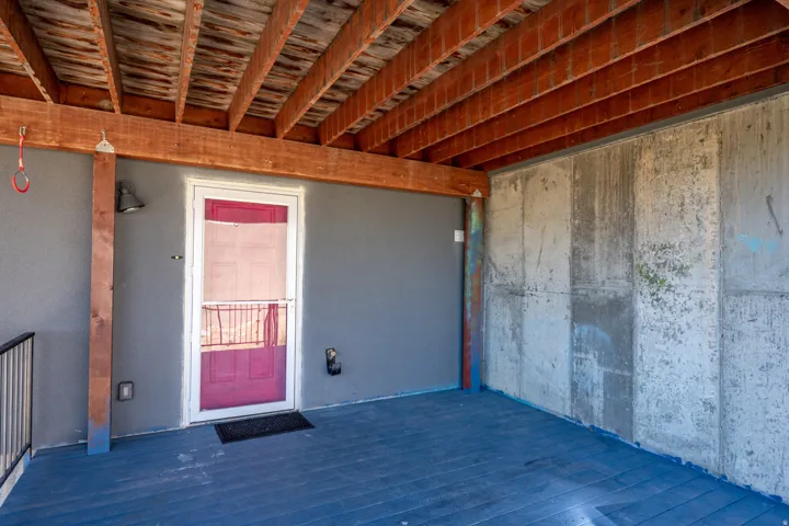 Covered outdoor space featuring a red entry door with a screen, exposed ceiling joists, and a concrete wall