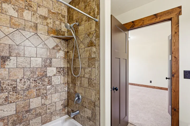 Bathtub shower featuring multicolored natural stone-look tiling with a decorative border, chrome fixtures, and an integrated corner shelf