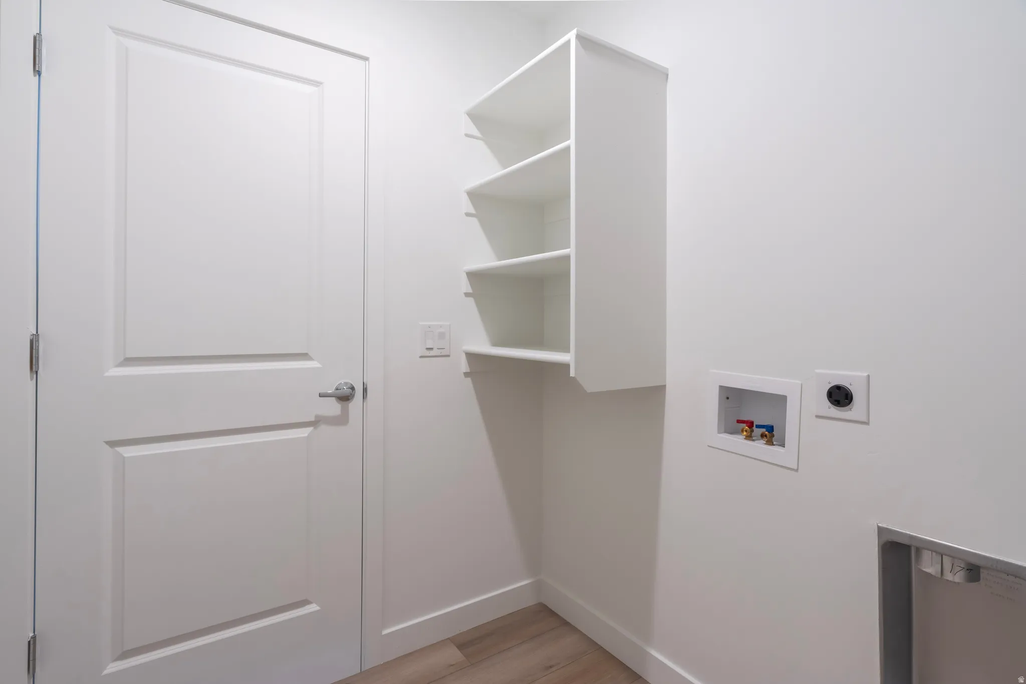 White paneled door with brushed nickel hardware, built-in white shelving, wood-finish flooring, and laundry hookups