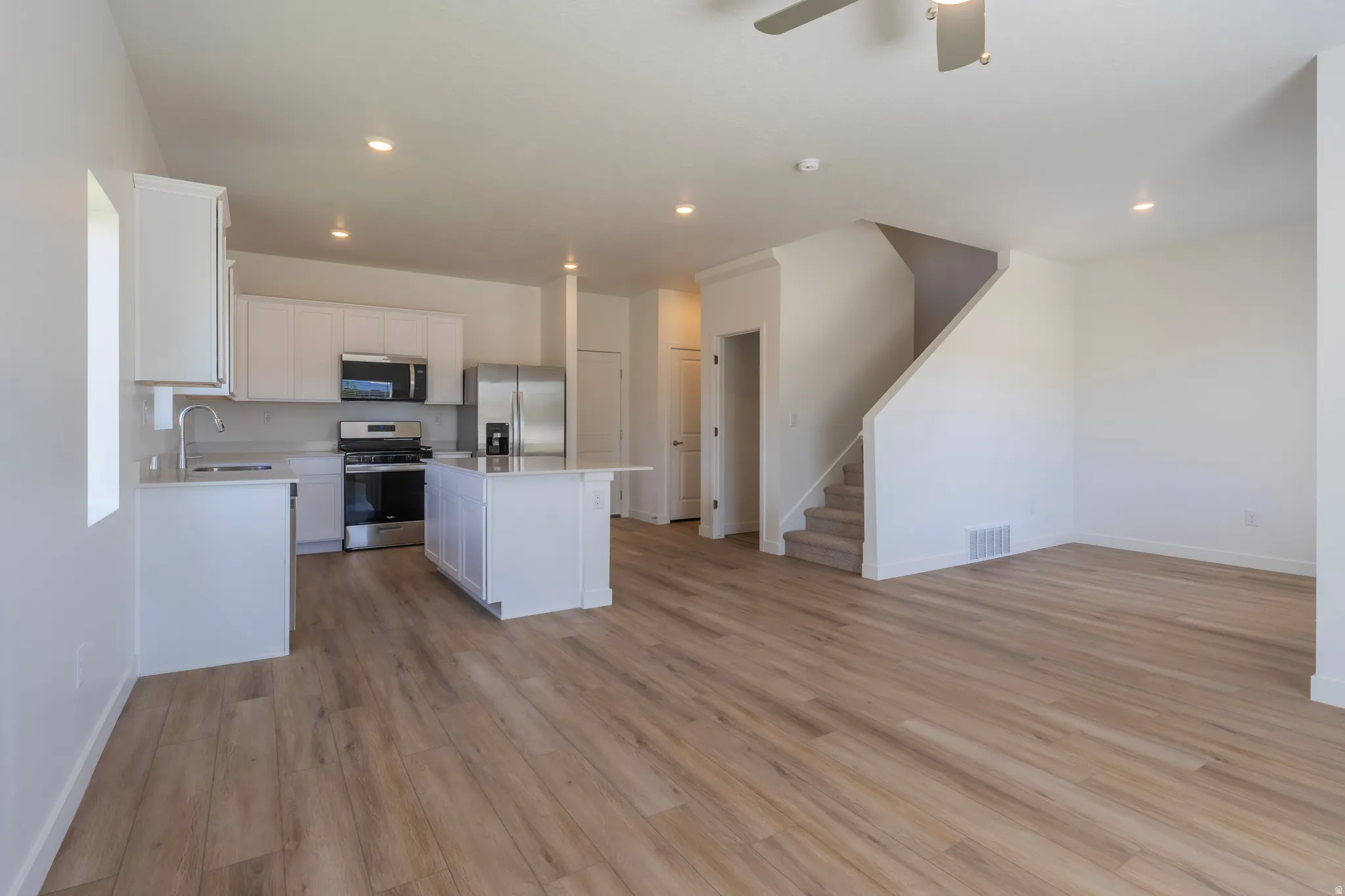 White kitchen cabinetry, stainless steel appliances, central kitchen island, wood-finish flooring, and a carpeted staircase