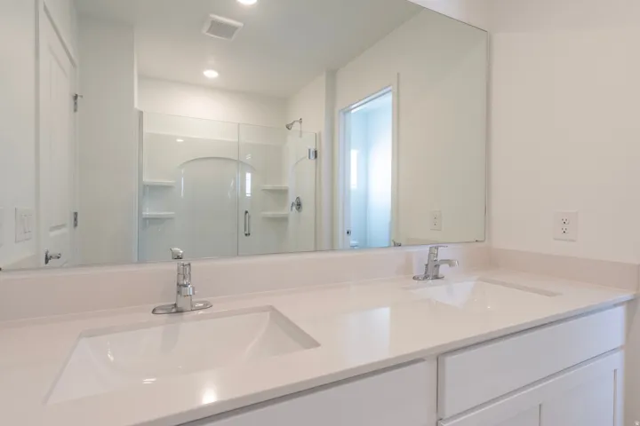 Dual undermount sinks with chrome faucets, light-toned countertop, white cabinetry, and a large wall mirror