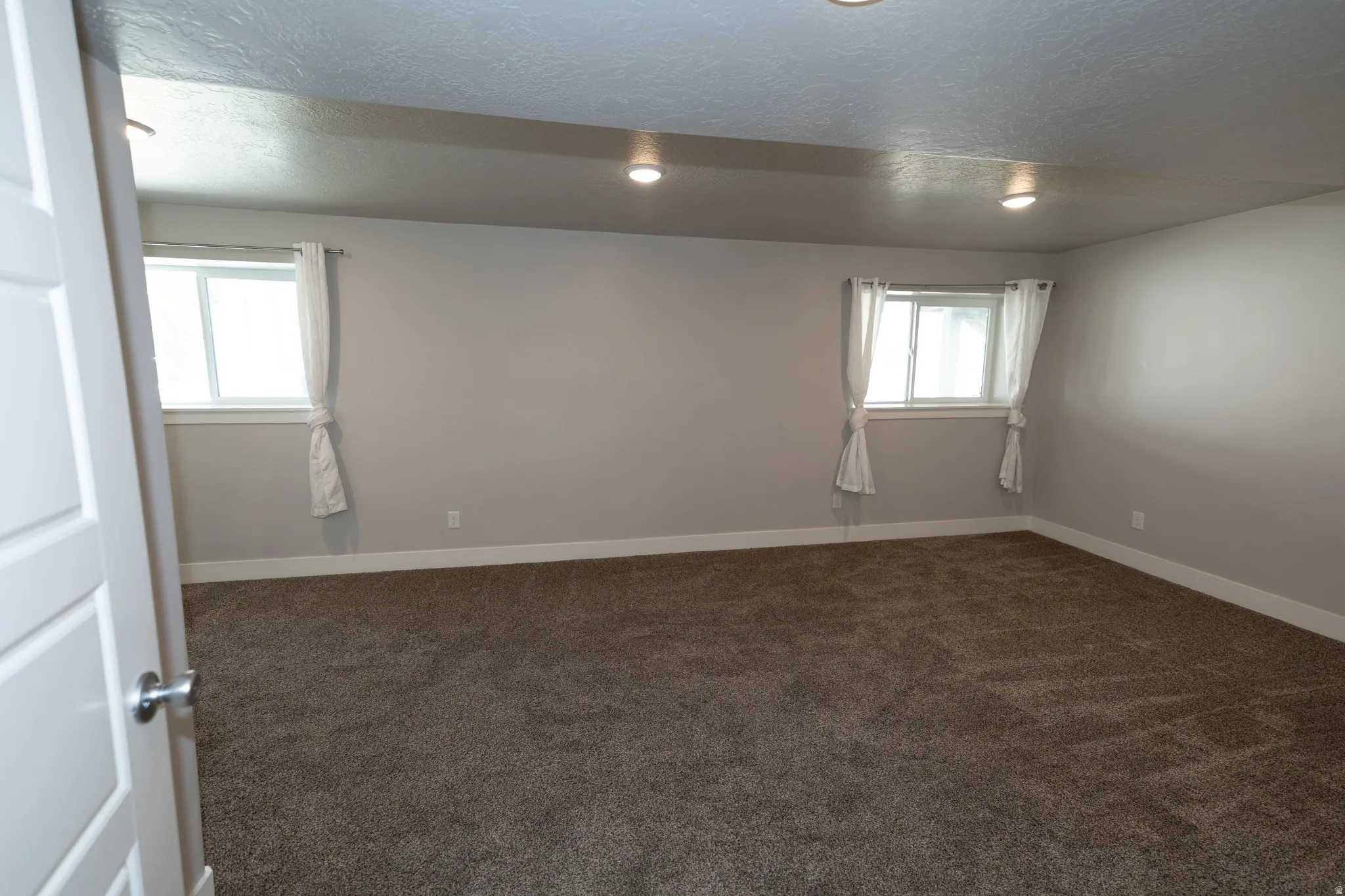 Spacious room featuring textured carpeting, neutral wall finishes, white trim, recessed lighting, and two windows with white curtains
