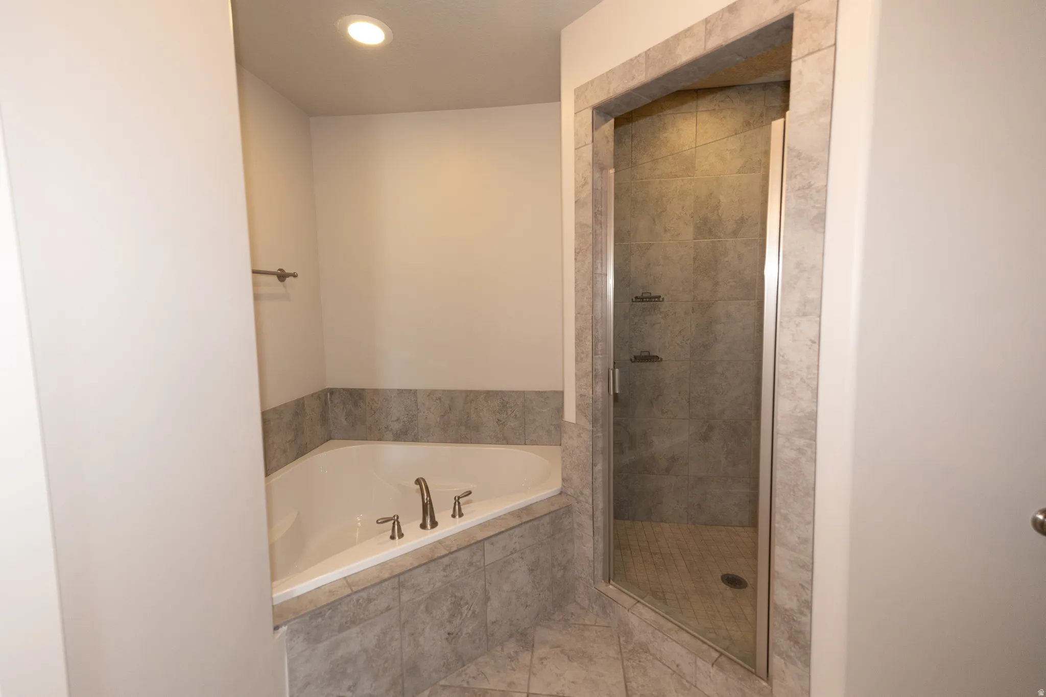 Corner soaking tub with deck-mounted brushed nickel fixtures, surrounded by large format tile, alongside a glass-enclosed shower with a tiled interior and floor, all set on coordinating tile flooring