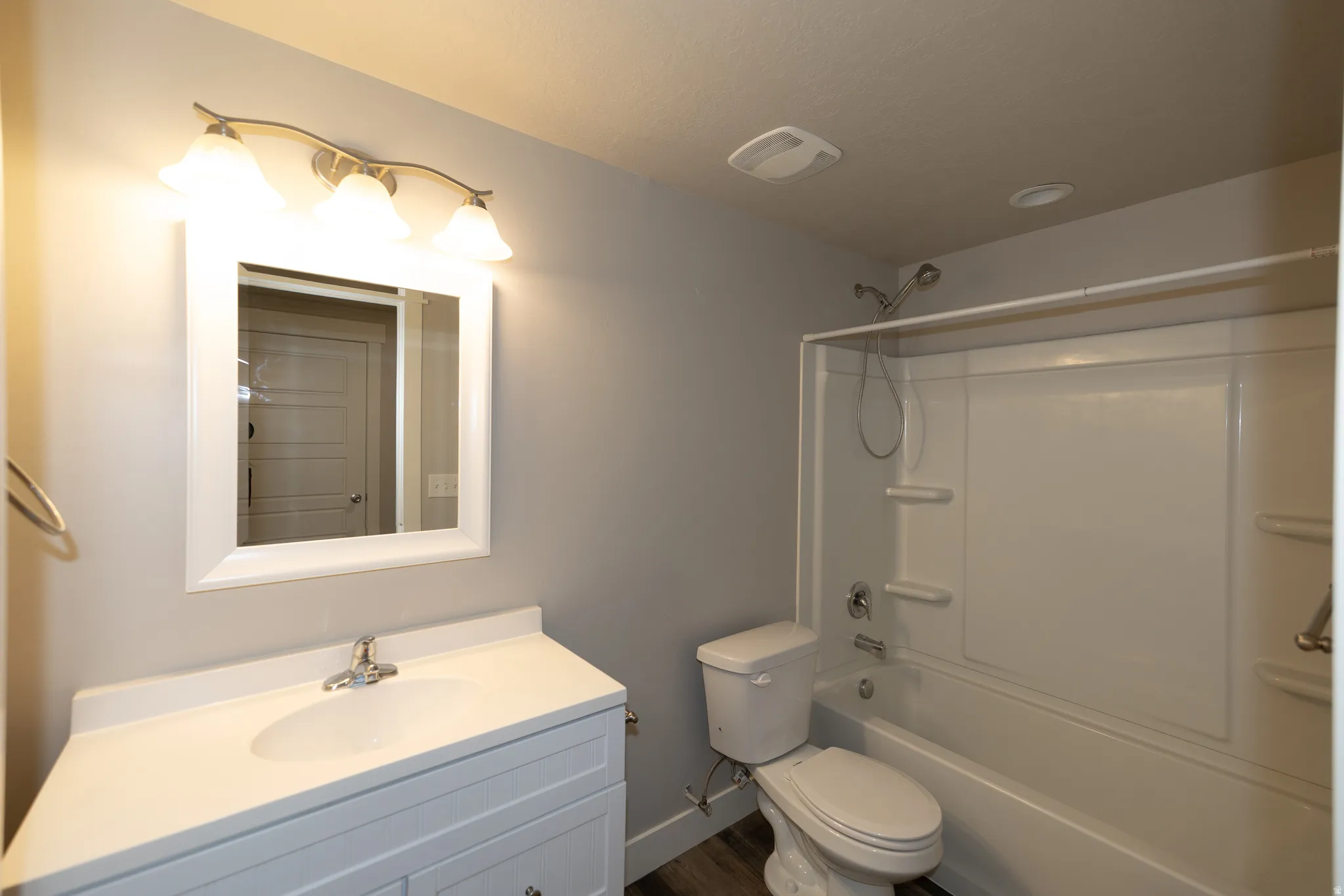 Bathroom featuring a single vanity with an integrated sink, a white framed mirror, a three-light vanity fixture, a white toilet, and a white tub/shower combination with a built-in shelf