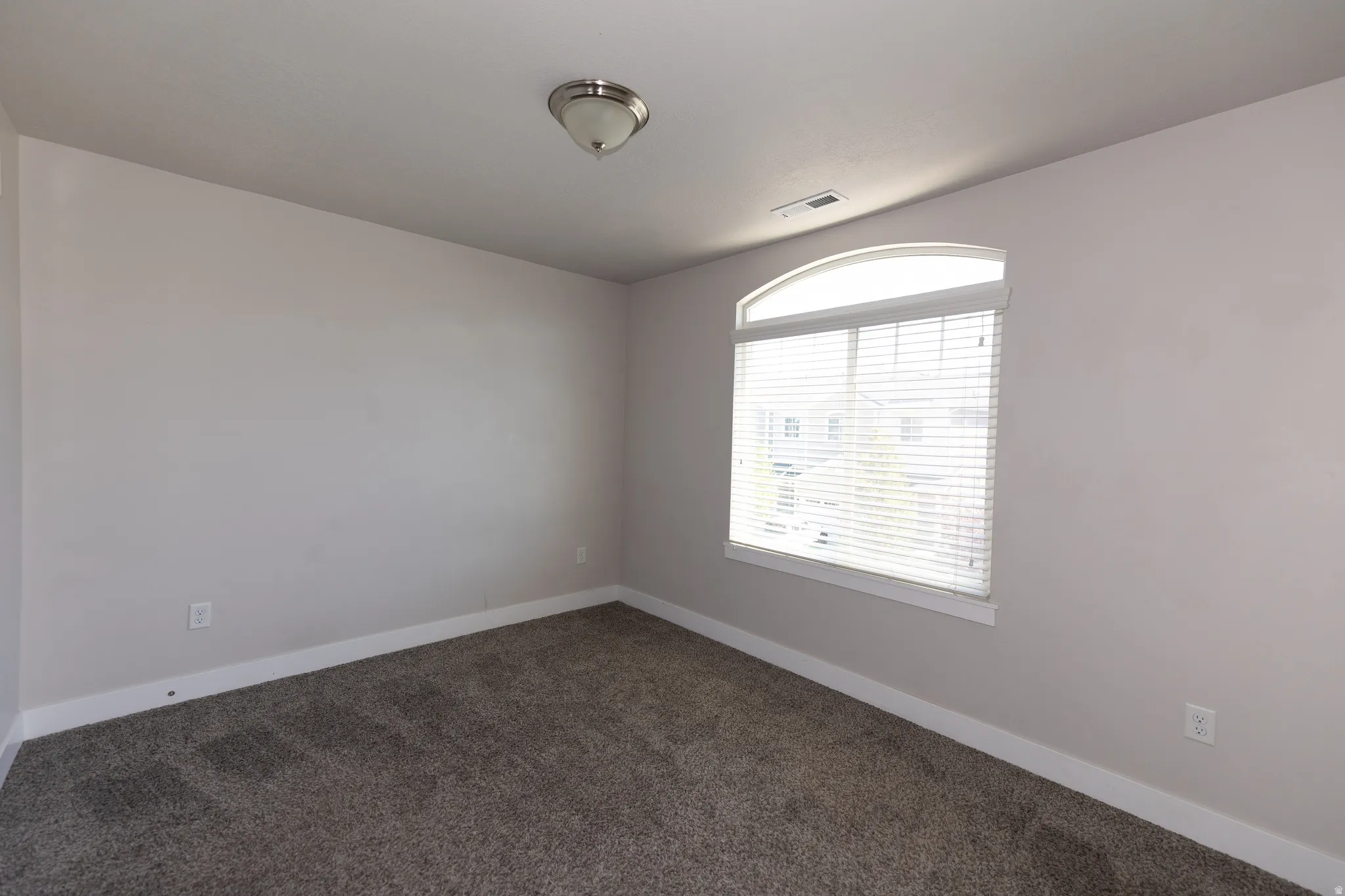 Room featuring neutral grey wall paint, plush grey carpeting, white baseboards, and a large arched window with horizontal blinds