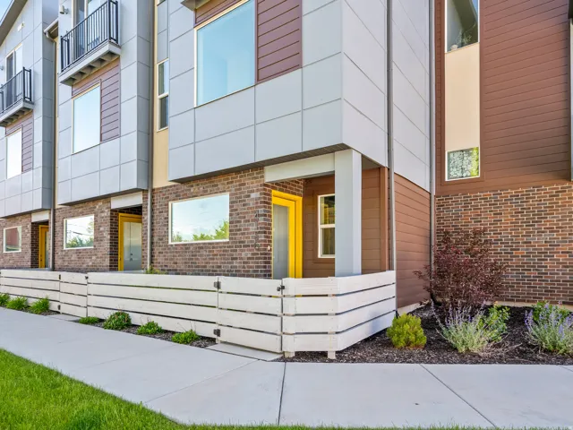 Contemporary townhome exterior featuring a white slatted privacy fence, brown brick facade, modern light gray paneling, and bright yellow entry door
