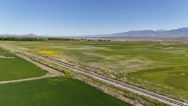 Expansive agricultural land featuring a railway line, vibrant green fields, distant mountains, and a clear blue sky