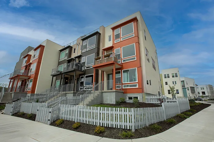 Modern townhome exteriors featuring varied siding materials and colors, contemporary window designs, multiple private balconies, individual front entrances with concrete steps, and white picket fences enclosing landscaped front yards