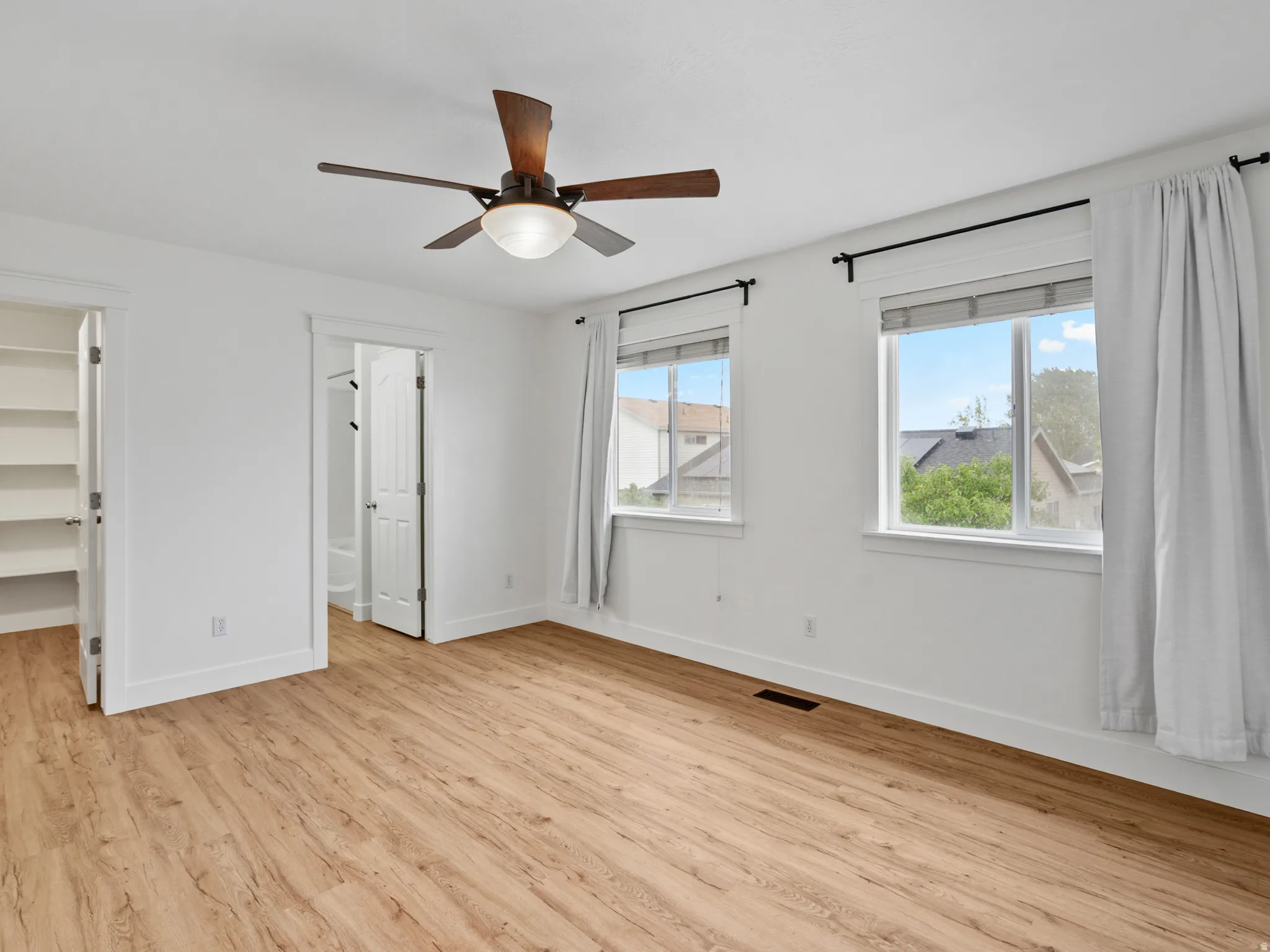 Spacious room featuring wood-finish flooring, two windows with white trim, a ceiling fan with integrated lighting, and a white interior door