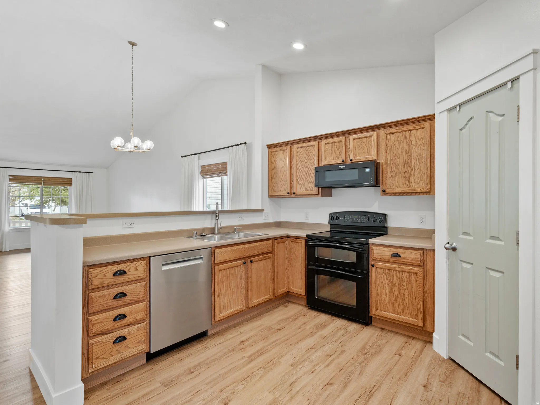 Open-concept kitchen featuring wood-finish flooring, light wood cabinetry, stainless steel sink, and black appliances