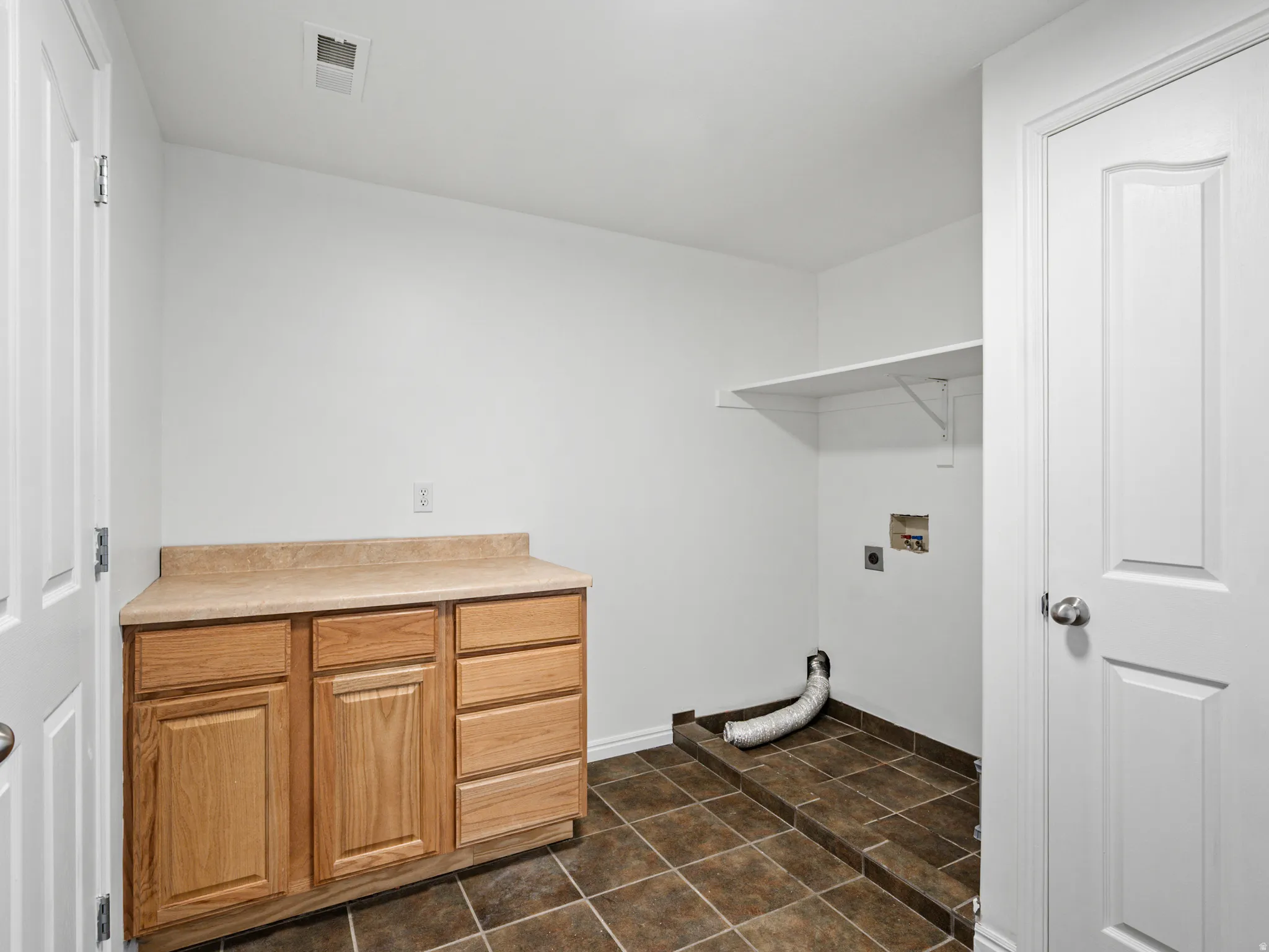 Utility room featuring a built-in wood-finish cabinet with a light-colored countertop, tiled flooring, and a wall-mounted shelving unit