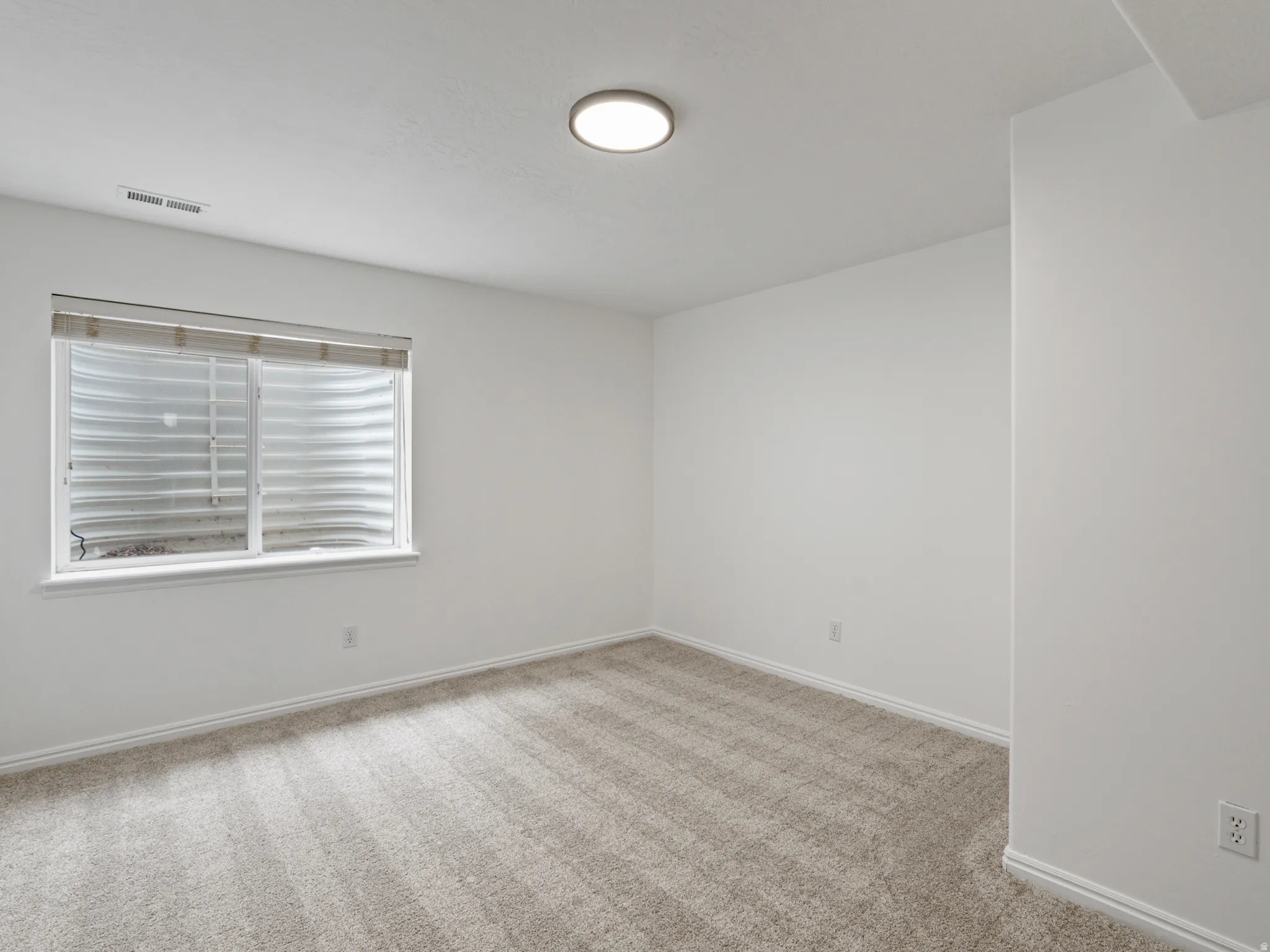 Recessed window with metal grate, light-toned carpet flooring, white baseboards, flush mount ceiling light, and white walls