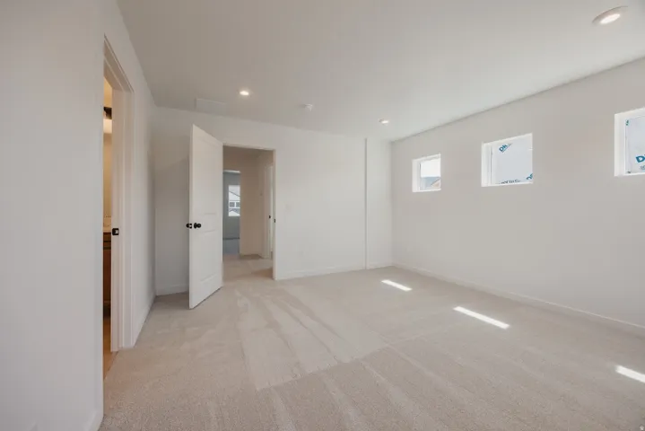 Open room with neutral carpeting, recessed ceiling lighting, three transom windows, a solid panel interior door with dark hardware, and a partial glimpse of a vanity with a light wood finish