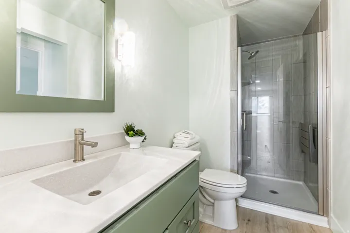 Bathroom featuring a glass-enclosed shower with tiled walls, a vanity with an integrated sink and light-colored countertop, green cabinetry, and a framed mirror with wall-mounted lighting
