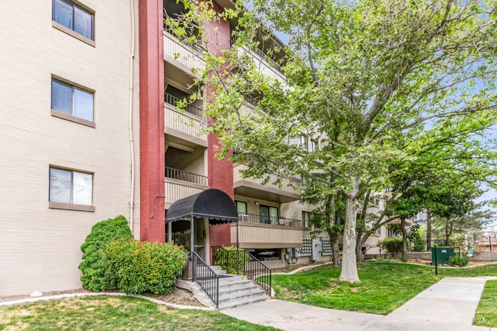 Multi-story building exterior featuring brick facade, private balconies, a covered entry with black awning, concrete steps with wrought iron railing, and established landscaping with mature trees