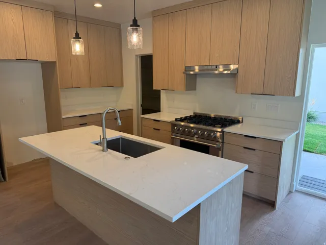 Kitchen featuring light wood-finish cabinetry, white countertops, a stainless steel gas range, a kitchen island with an undermount sink, and pendant lighting