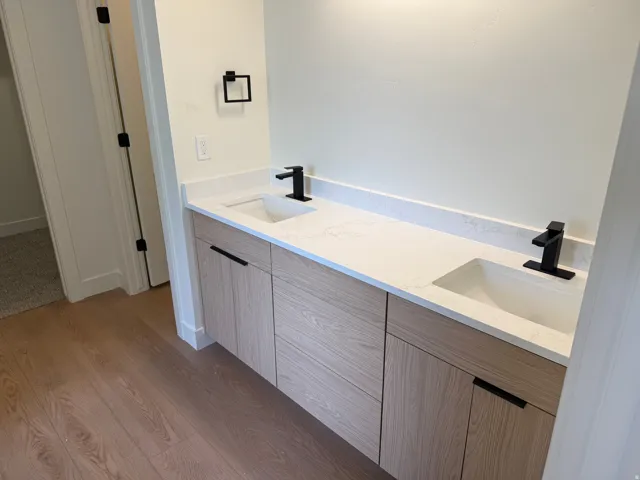 Dual vanity with integrated sinks, light wood-finish cabinetry, matte black hardware, and stone-look countertops