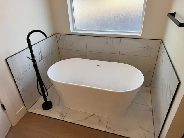 Freestanding soaking tub with a matte black floor-mounted faucet, complemented by marble-look tile flooring and wainscoting, and a frosted privacy window