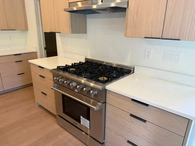 Stainless steel range with a ventilation hood, light wood-finish cabinetry, white countertops, and wood-finish flooring