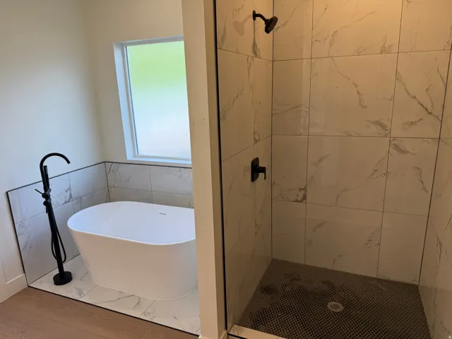 Freestanding soaking tub with a floor-mounted matte black faucet, tiled shower with glass enclosure and black fixtures, and wood-finish flooring