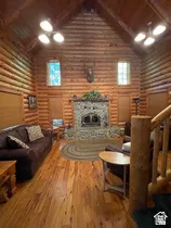 Vaulted ceiling great room featuring log walls, wood-finish flooring, and a stone fireplace with a wood-burning insert