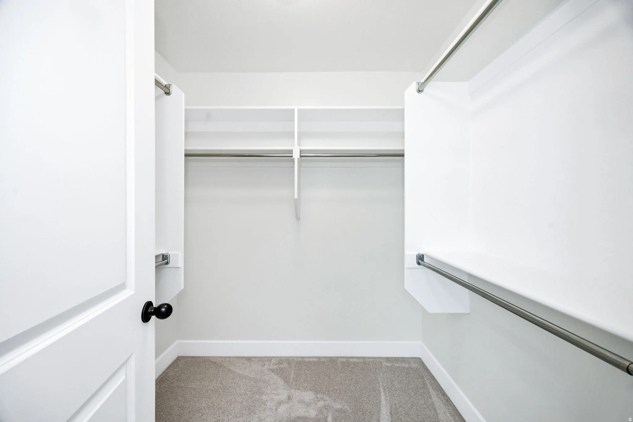 Spacious walk-in closet featuring white built-in shelving, chrome hanging rods, and light gray carpet flooring