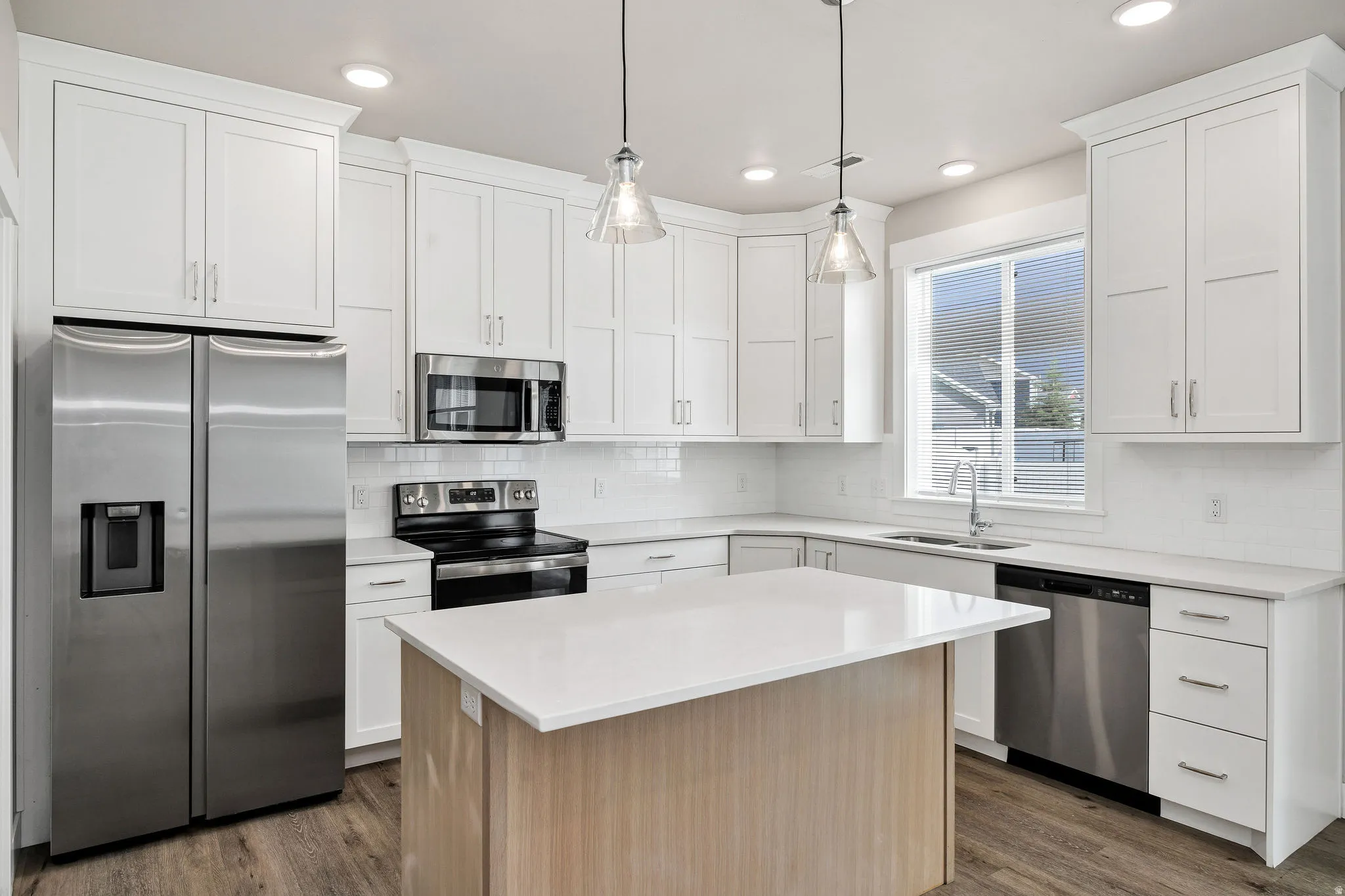 Kitchen featuring white cabinetry with brushed nickel hardware, stainless steel appliances, a center island with a light wood-finish base and light-toned countertop, pendant lighting, and wood-finish flooring