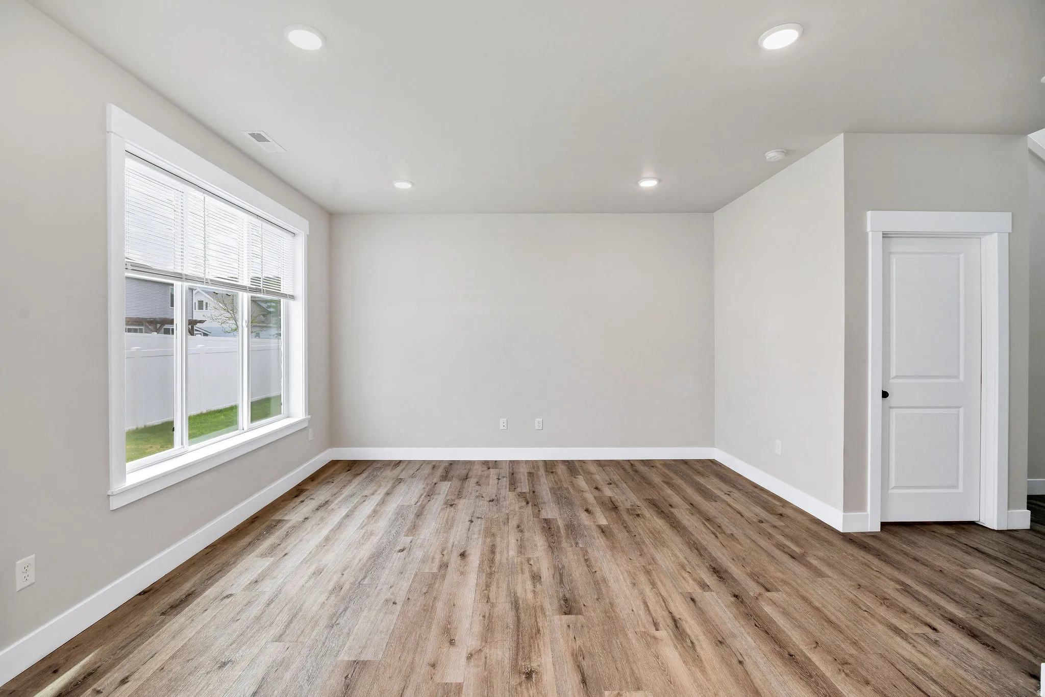 Spacious room featuring wood-finish flooring, a large multi-pane window with blinds, recessed ceiling lighting, and a paneled interior door with black hardware