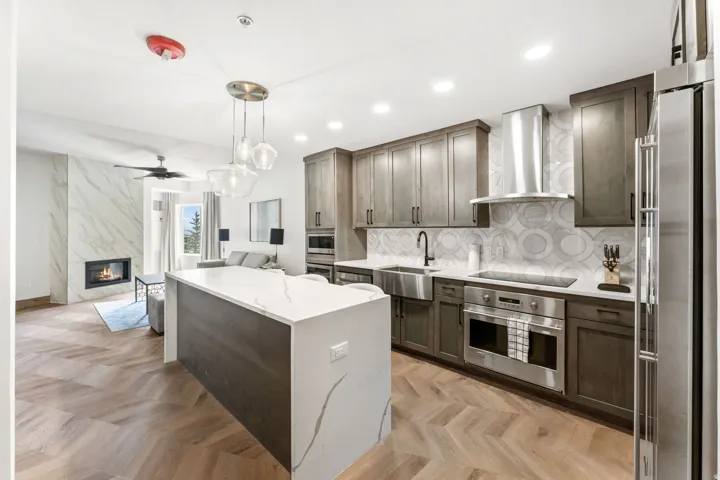 Open-concept living space featuring a herringbone wood-finish floor, a central kitchen island with a white countertop, dark wood-finish cabinetry, stainless steel appliances, and a marble-clad fireplace