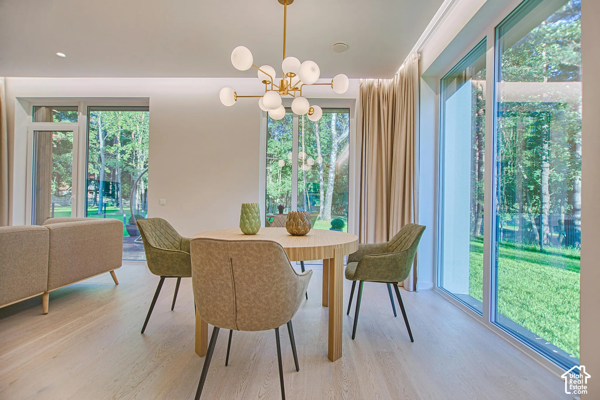 Light-filled interior featuring floor-to-ceiling glass windows, wide-plank wood-finish flooring, a multi-arm modern brass chandelier, and recessed ceiling lighting