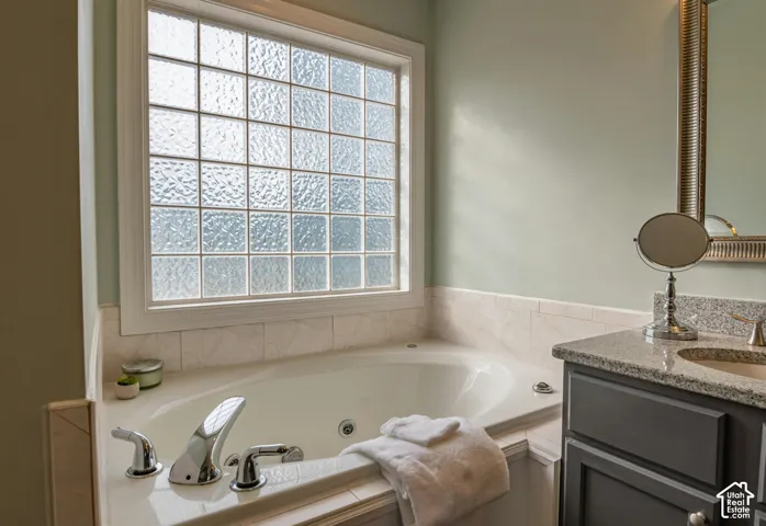 Built-in soaking tub with chrome fixtures, grid-pattern textured glass window, and vanity featuring a stone countertop