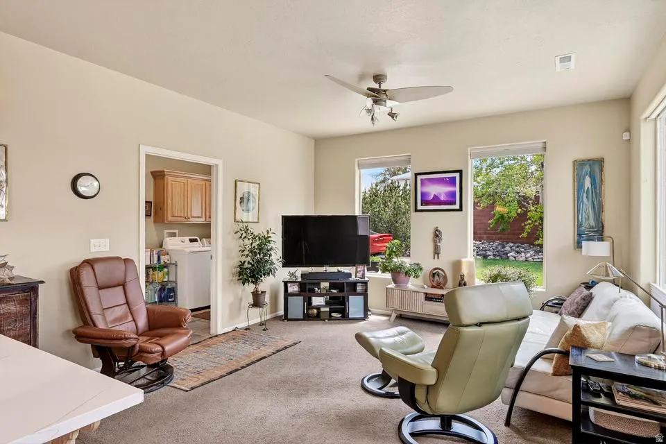 Carpeted living area featuring multiple windows, a ceiling fan with light fixture, and a doorway leading to a utility space with wood cabinetry and a washer